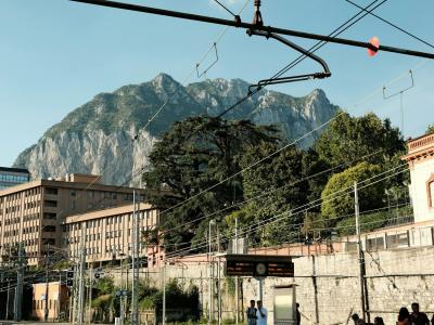 A view of a large, rocky mountain peak rising behind urban buildings and railway overhead lines, with a sign reading "GROSTÈ" visible on a structure in the foreground.