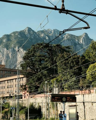 A view of a large, rocky mountain peak rising behind urban buildings and railway overhead lines, with a sign reading "GROSTÈ" visible on a structure in the foreground.