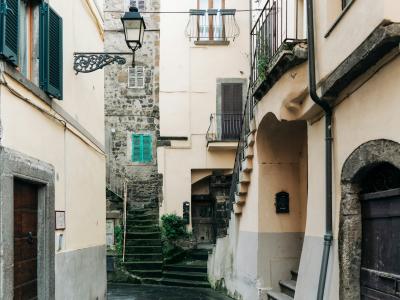 A narrow, steep cobblestone alleyway in a historic Italian town, featuring stone buildings with shuttered windows, an external staircase, and a vintage-style street lamp.