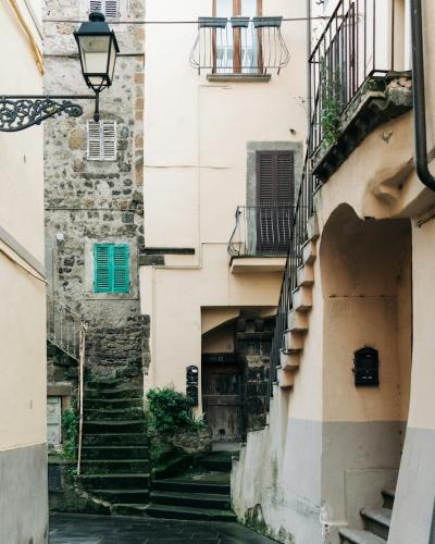 A narrow, steep cobblestone alleyway in a historic Italian town, featuring stone buildings with shuttered windows, an external staircase, and a vintage-style street lamp.