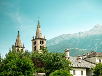 Two stone church towers with pointed spires and crosses, set against a backdrop of green trees and a large mountain under a blue sky.