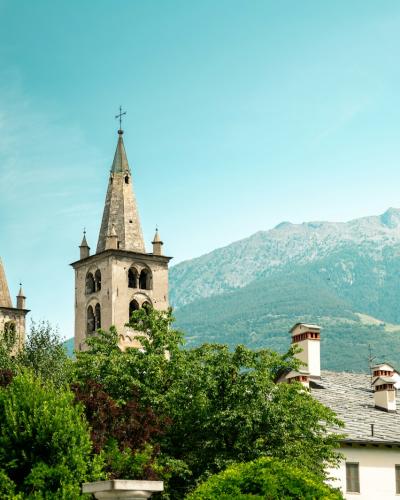 Two stone church towers with pointed spires and crosses, set against a backdrop of green trees and a large mountain under a blue sky.