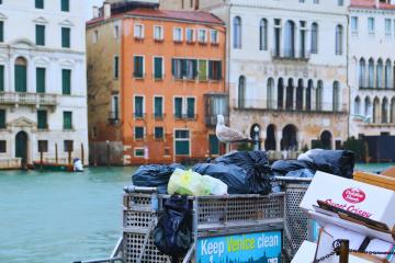 A barge on a canal in Venice, Italy, piled with black trash bags and a white recycling bin, with a seagull perched on the bags and a sign reading "Keep Venice clean" visible on the side of the barge.