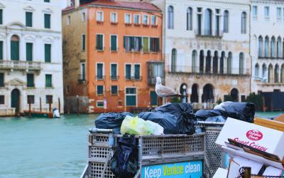 A barge on a canal in Venice, Italy, piled with black trash bags and a white recycling bin, with a seagull perched on the bags and a sign reading "Keep Venice clean" visible on the side of the barge.