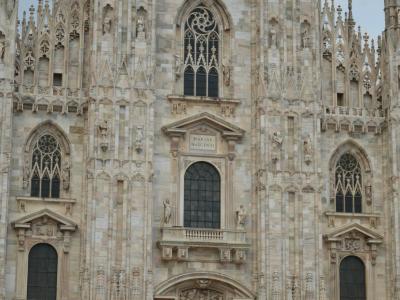 The image shows the ornate Gothic facade of the Milan Cathedral (Duomo di Milano), featuring pointed arches, statues, and intricate stone carvings under daylight.