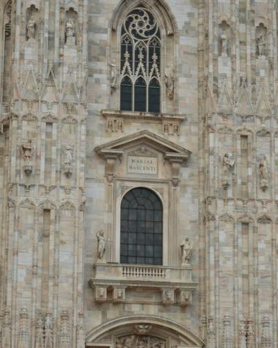 The image shows the ornate Gothic facade of the Milan Cathedral (Duomo di Milano), featuring pointed arches, statues, and intricate stone carvings under daylight.
