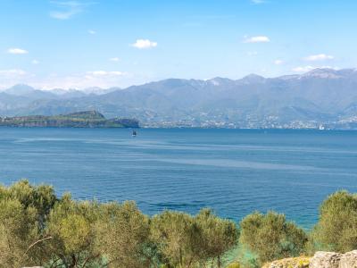 A scenic view of Lake Garda framed by ancient stone ruins in the foreground, with distant mountains and a clear blue sky.