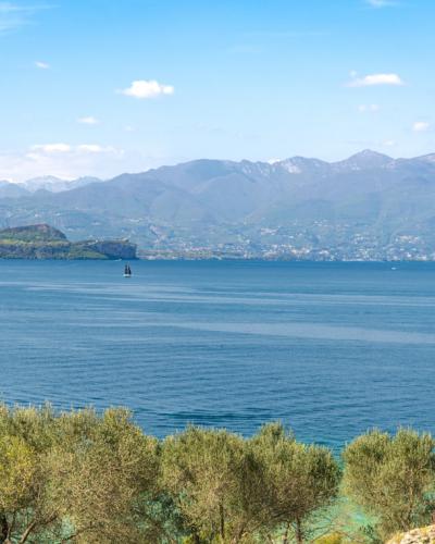 A scenic view of Lake Garda framed by ancient stone ruins in the foreground, with distant mountains and a clear blue sky.