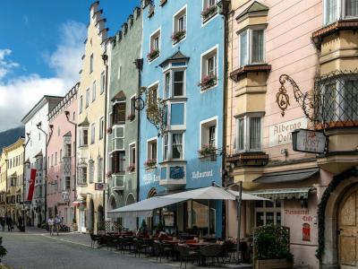 A row of colorful, multi-story historic buildings with stepped gables and ornate facades lines a paved pedestrian street under a blue sky, with outdoor café seating in the foreground and mountains visible in the distance.