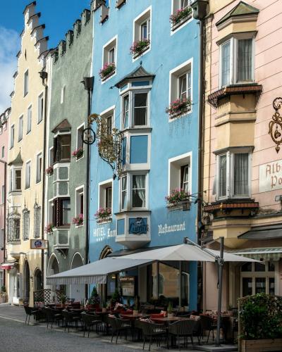 A row of colorful, multi-story historic buildings with stepped gables and ornate facades lines a paved pedestrian street under a blue sky, with outdoor café seating in the foreground and mountains visible in the distance.