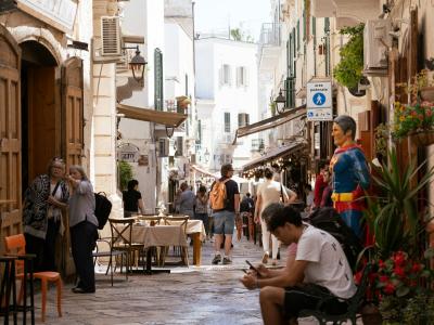 A narrow, sunlit pedestrian street in a historic European town, featuring stone-paved ground, outdoor café seating, and a prominent Superman statue on the right side.