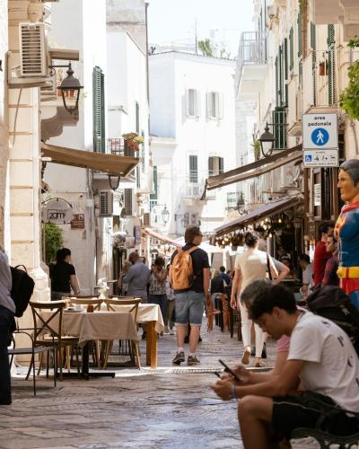 A narrow, sunlit pedestrian street in a historic European town, featuring stone-paved ground, outdoor café seating, and a prominent Superman statue on the right side.