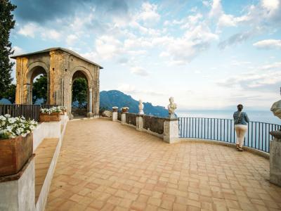 A terraced stone terrace with Moorish-style arches, white statues, and a woman looking out over the sea towards mountains in the distance.
