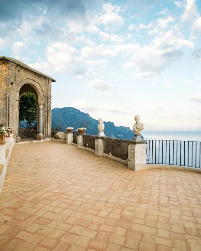 A terraced stone terrace with Moorish-style arches, white statues, and a woman looking out over the sea towards mountains in the distance.
