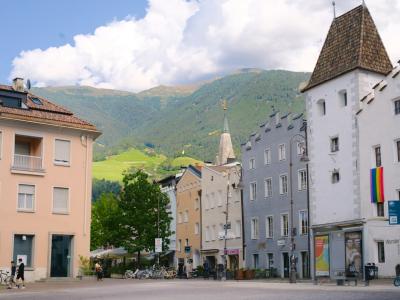A street scene in a European town featuring colorful buildings, a prominent white tower with a flag, and mountains in the background under a cloudy sky.