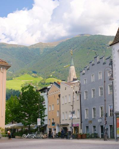 A street scene in a European town featuring colorful buildings, a prominent white tower with a flag, and mountains in the background under a cloudy sky.