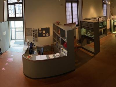 The image shows the interior entrance and reception area of a museum, featuring a curved service desk, display cases with artifacts, informational signage, and an open door leading outside.