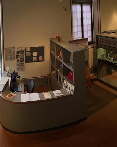 The image shows the interior entrance and reception area of a museum, featuring a curved service desk, display cases with artifacts, informational signage, and an open door leading outside.