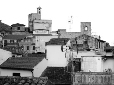 A black-and-white photograph showing a dense cluster of terracotta-tiled roofs, whitewashed walls, and prominent stone structures including a castle tower and a bell tower, with a hillside visible in the background.