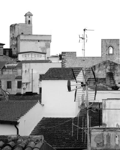 A black-and-white photograph showing a dense cluster of terracotta-tiled roofs, whitewashed walls, and prominent stone structures including a castle tower and a bell tower, with a hillside visible in the background.