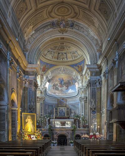 The interior of a grand Baroque-style church featuring marble columns, an ornate vaulted ceiling, a large apse painting, and a prominent golden statue of the Virgin Mary on the left side.