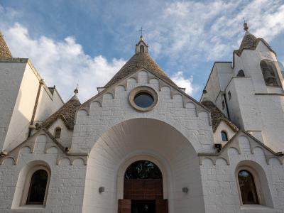 A low-angle photograph of a white stone church with multiple conical roofs and a central arched entrance, set against a blue sky with clouds.
