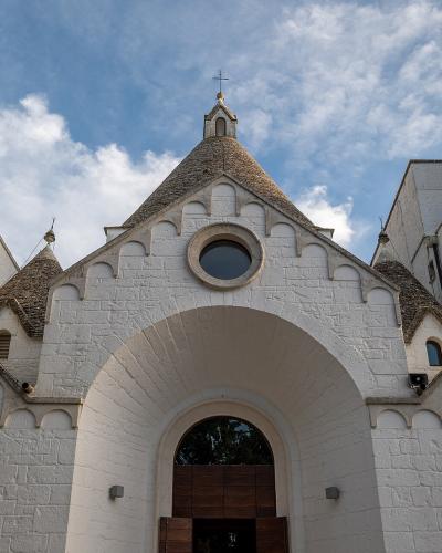 A low-angle photograph of a white stone church with multiple conical roofs and a central arched entrance, set against a blue sky with clouds.