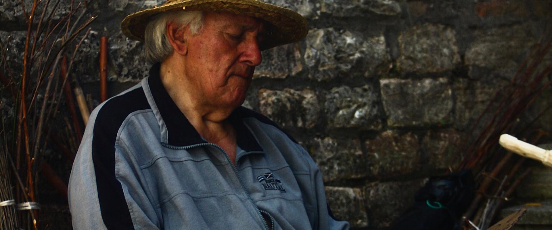 An elderly man wearing a straw hat and a grey tracksuit sits on a wooden chair against a rough stone wall, holding scissors and a piece of wood or cardboard, with bundles of thin branches nearby.