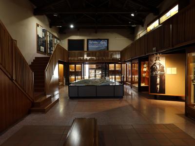 A museum interior featuring a large architectural model of an ancient site on a central pedestal, surrounded by glass display cases, wooden paneling, and a staircase.