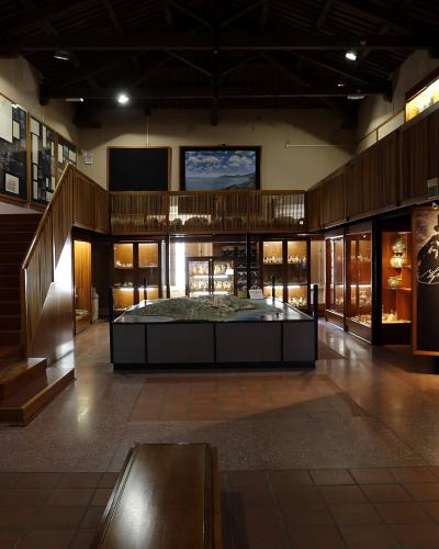 A museum interior featuring a large architectural model of an ancient site on a central pedestal, surrounded by glass display cases, wooden paneling, and a staircase.