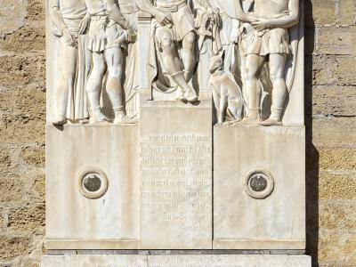 A stone bas-relief monument mounted on a yellowish-brown brick wall, featuring a central crowned figure seated on a throne flanked by standing figures and a dog, with Latin inscriptions below and two lion-head corbels supporting the base; text inc