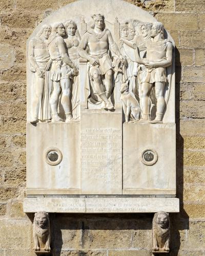 A stone bas-relief monument mounted on a yellowish-brown brick wall, featuring a central crowned figure seated on a throne flanked by standing figures and a dog, with Latin inscriptions below and two lion-head corbels supporting the base; text inc