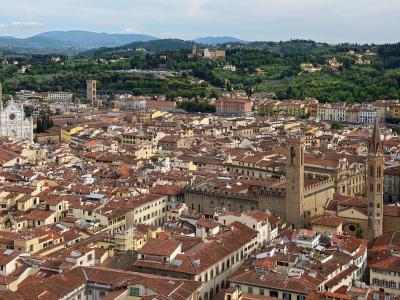 A panoramic high-angle view of Florence, Italy, showing the terracotta rooftops of the historic center, with the distinctive white marble facade of the Basilica of Santa Croce visible on the left and Giotto’s Campanile (bell tower) of the Duomo in