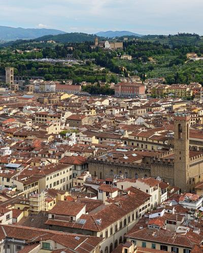 A panoramic high-angle view of Florence, Italy, showing the terracotta rooftops of the historic center, with the distinctive white marble facade of the Basilica of Santa Croce visible on the left and Giotto’s Campanile (bell tower) of the Duomo in