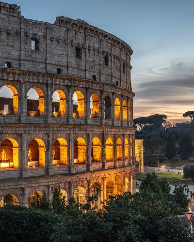 The image shows the exterior of the Colosseum in Rome, Italy, illuminated at dusk, with visible arches, ancient stonework, and signage reading "the Colosseum" inside one of the lower arches; the Roman Forum and surrounding archaeological landscape