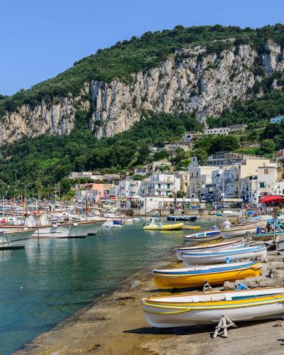 A sunlit harbor scene featuring numerous small boats moored in turquoise water, with a steep, rocky cliff face rising behind white buildings and a stone pier in the foreground where people sit and more boats are pulled ashore.