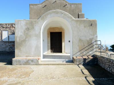 A small, whitewashed chapel with a prominent arched entrance and a simple gabled facade, set against a clear blue sky with stone walls and a metal railing visible to the right.