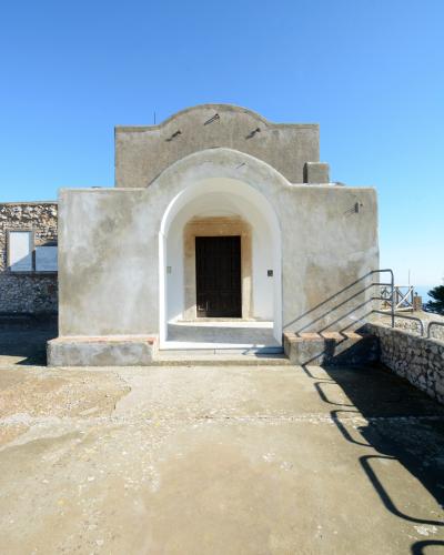 A small, whitewashed chapel with a prominent arched entrance and a simple gabled facade, set against a clear blue sky with stone walls and a metal railing visible to the right.