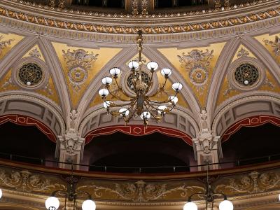 The image shows the ornate interior ceiling and upper balcony boxes of a historic opera house, featuring gold-leafed plasterwork, decorative medallions, arched box fronts with red valances, and large crystal chandeliers.