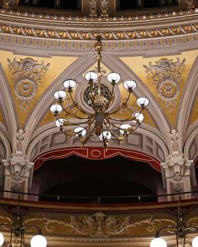 The image shows the ornate interior ceiling and upper balcony boxes of a historic opera house, featuring gold-leafed plasterwork, decorative medallions, arched box fronts with red valances, and large crystal chandeliers.