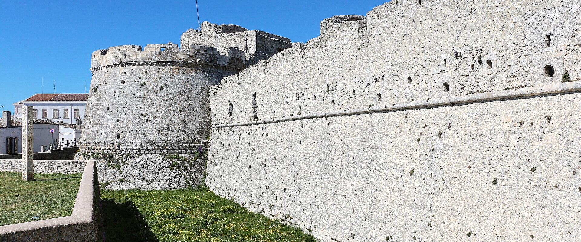 A low-angle view of a massive, light-colored stone fortress wall with a prominent round tower, set against a clear blue sky with a grassy foreground and a white building visible in the background.