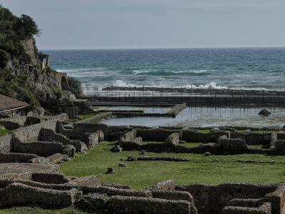 The image shows the archaeological ruins of the Villa of Tiberius at Sperlonga, featuring low stone walls, a grassy area, and a fenced-off section near the sea with a cliff face on the left.