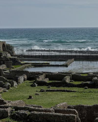The image shows the archaeological ruins of the Villa of Tiberius at Sperlonga, featuring low stone walls, a grassy area, and a fenced-off section near the sea with a cliff face on the left.
