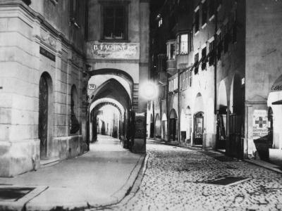 A black-and-white photograph of a narrow, cobblestone street in an old European city, featuring a prominent arched passageway on the left with a sign reading "B. FACHINETTI Fabbrica" and another sign for "Kofler" on the right.