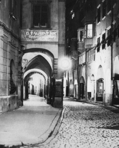 A black-and-white photograph of a narrow, cobblestone street in an old European city, featuring a prominent arched passageway on the left with a sign reading "B. FACHINETTI Fabbrica" and another sign for "Kofler" on the right.