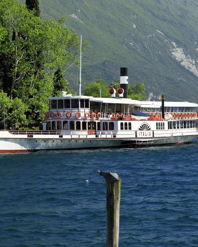 A white and red paddle steamer-style boat with "ITALIA" written on its side is docked or moving slowly near a tree-lined shore with a large mountain in the background; people are visible on a pier to the left.