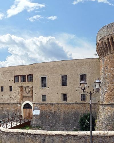 A large, historic stone fortress built atop a steep, rocky tidal island rising directly from the sea, with small boats in the foreground water and a blue sky with wispy clouds above.