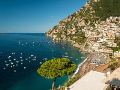 A high-angle view of the coastal village of Positano on the Amalfi Coast, Italy, showing its colorful buildings cascading down a steep cliffside to a pebble beach lined with umbrellas, with numerous boats anchored in the turquoise bay.