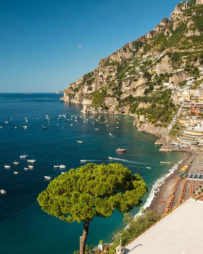 A high-angle view of the coastal village of Positano on the Amalfi Coast, Italy, showing its colorful buildings cascading down a steep cliffside to a pebble beach lined with umbrellas, with numerous boats anchored in the turquoise bay.