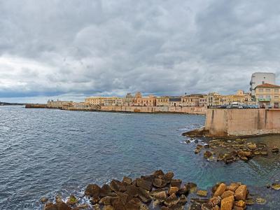 A coastal view of a historic walled city with ochre and yellow buildings lining the seafront under a cloudy sky, featuring a stone breakwater and rocky shoreline in the foreground.